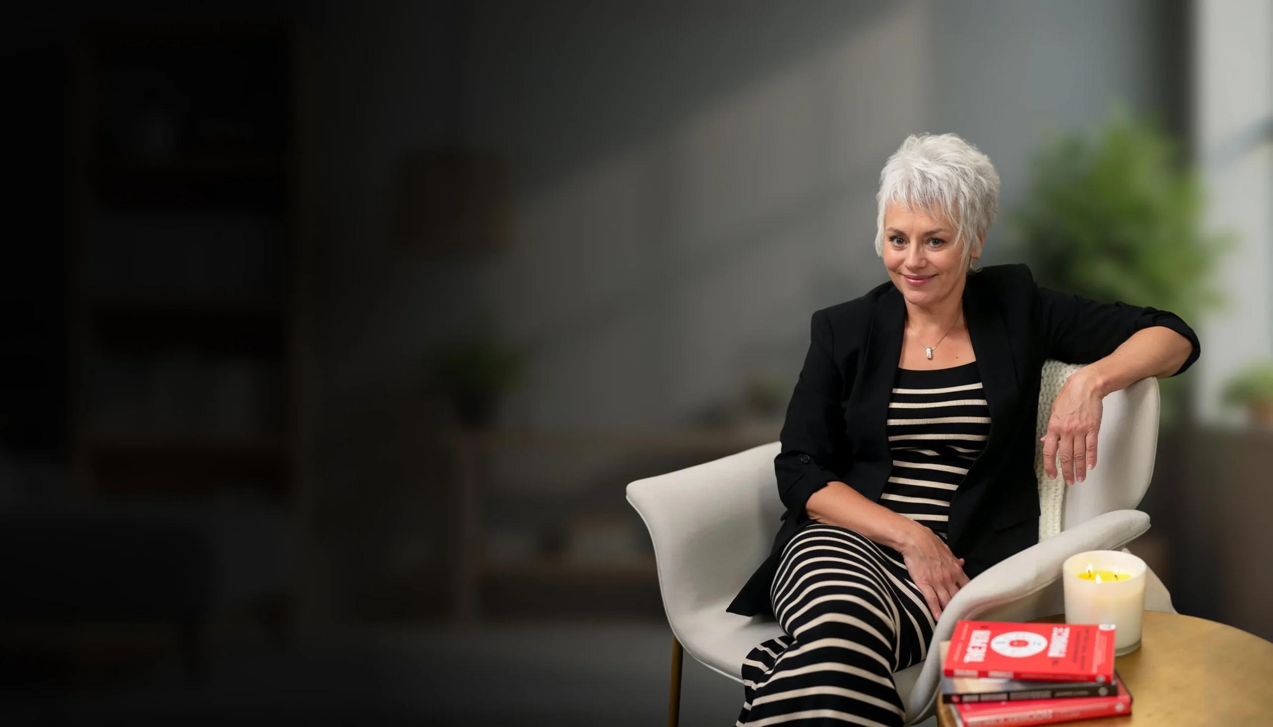Jackie Vick sitting on a chair in a modern office, wearing a black blazer and striped dress, with a confident smile